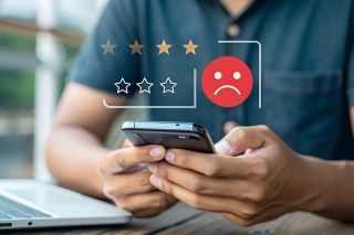 © main_asn - stock.adobe.com Foto: Mann bedient Smartphone, darüber negatives Smiley und schlechte Sterne Bewertung